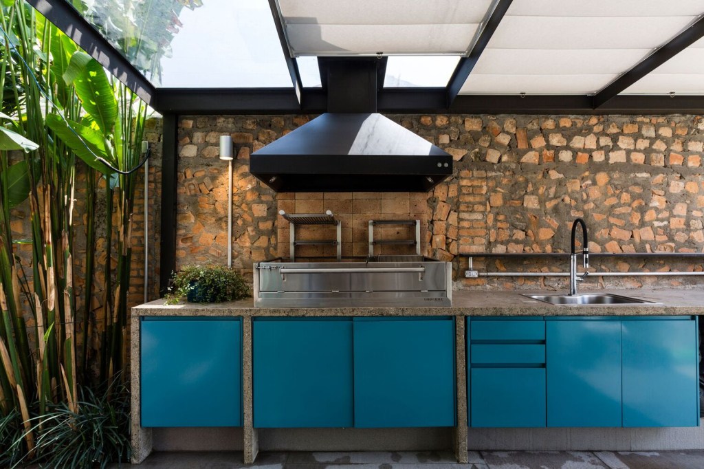 Área gourmet tem parede de entulho na bancada de trabalho com armários pintados de azul e cobertura translúcida integrada ao jardim.