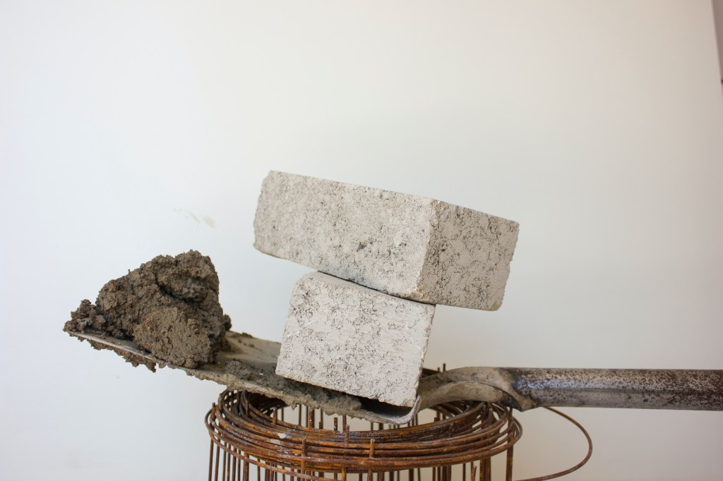 Sobre fundo branco, vemos restos de materiais de construção: vergalhão em malha, pá com cimento e dois blocos de pedra.