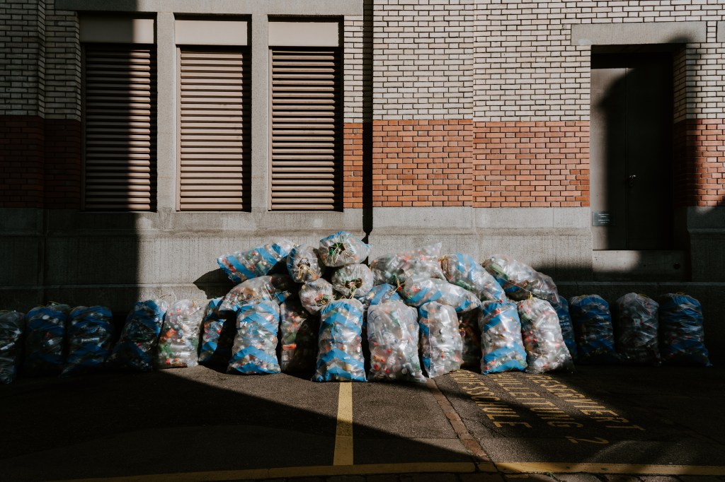 Imagem mostra dezenas de sacos plásticos com restos de materiais colocados na frente de uma casa em que vemos a fachada de tijolos e parte de uma janela.
