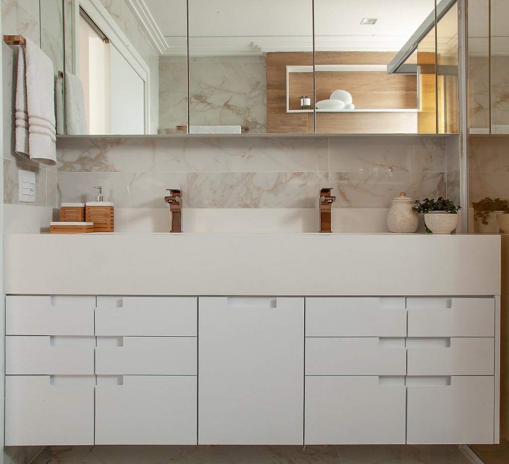 Bancada de banheiro com marcenaria branca com gavetas, duas torneiras de mesa em tom de cobre, espelho tomando toda a parede e tomada na parede lateral esquerda.