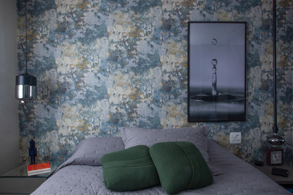 Quarto com cama de casal com colcha cinza e duas almofadas verdes e parede de cabeceira com papel de parede floral em tons de azul e bege, com quadro de gota d'água à direita e tomada ao lado da cabeceira.