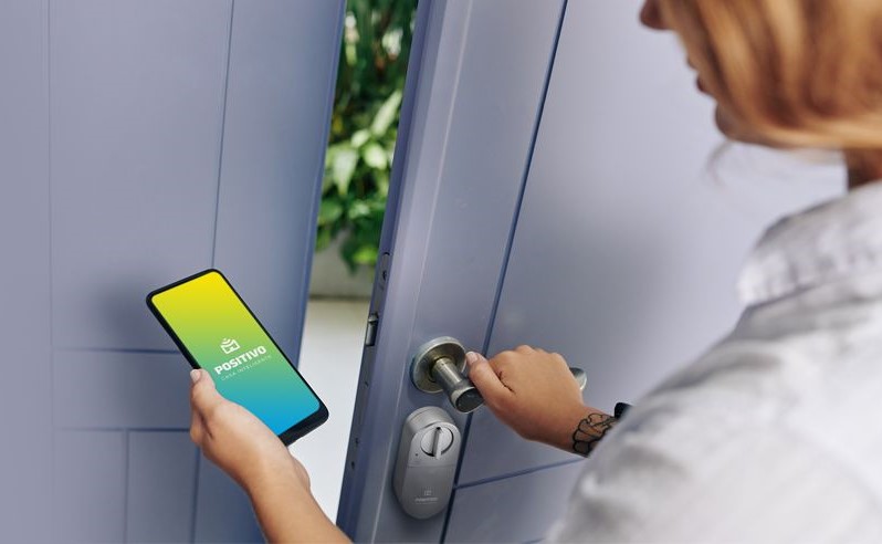 Pessoal abre porta enquanto segura um celular cuja tela mostra o aplicativo da Positivo Casa Inteligente.
