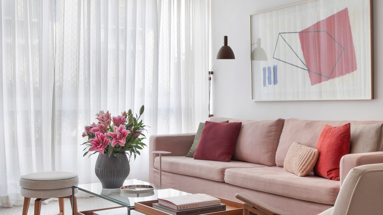 Sala de estar com sofá rosa e mesa de centro. Cortina branca ao fundo