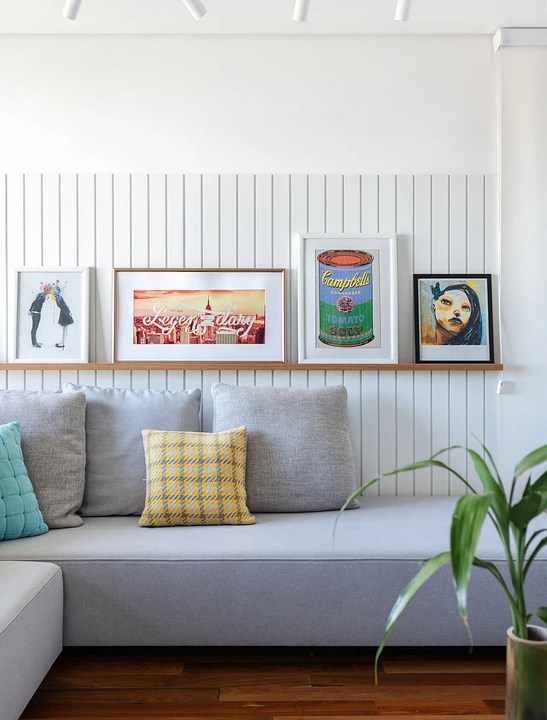 Sala de estar com sofá cinza; parede em madeira ripada; quadros em prateleira.