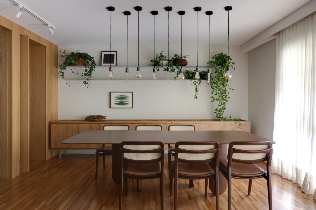 Sala de jantar com mesa de seis lugares; buffet ao fundo; luminárias pendentes e prateleiras com plantas