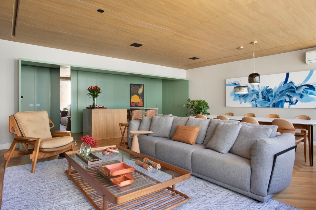 Sala de tv com rack em madeira, sofá cinza, tapete, estante em madeira com prateleiras e mesa de centro