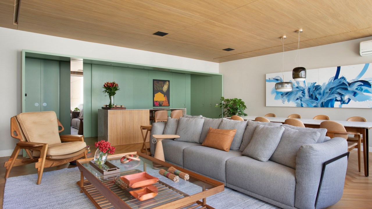 Sala de tv com rack em madeira, sofá cinza, tapete, estante em madeira com prateleiras e mesa de centro