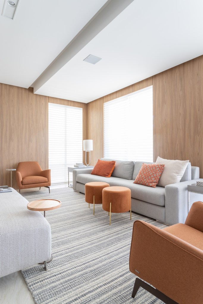 Sala de estar; paredes revestidas de madeira; sofá cinza; poltrona laranja; almofadas laranjas; tapete
