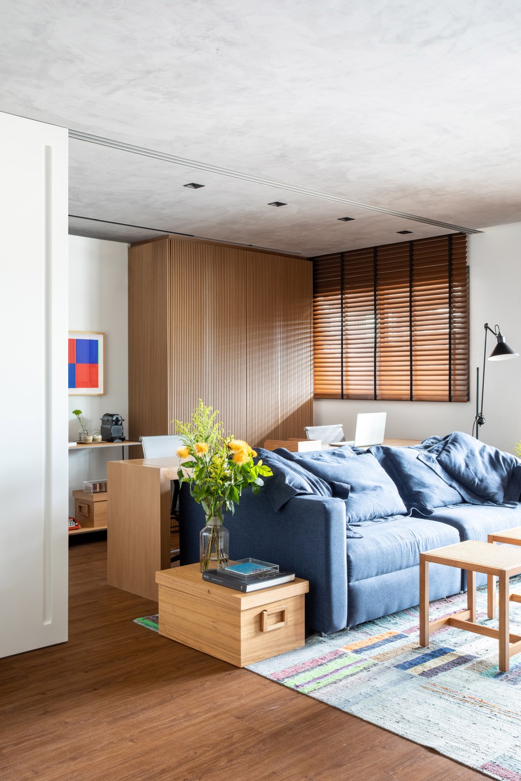 Sala de estar; sala de estar com home office; bancada de home office; persiana de madeira; sofá azul; tapete azul.