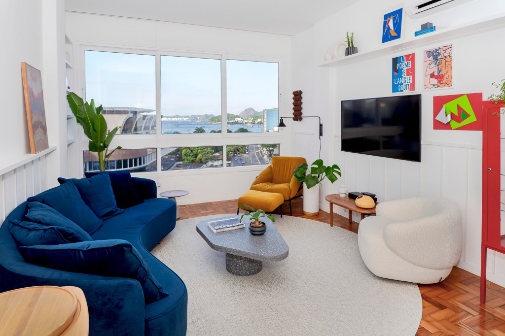 Sala de estar; sala de estar clara; sofá azul marinho; sofá curvo; poltrona branca; poltrona mostarda; tapete branco. mesa de centro