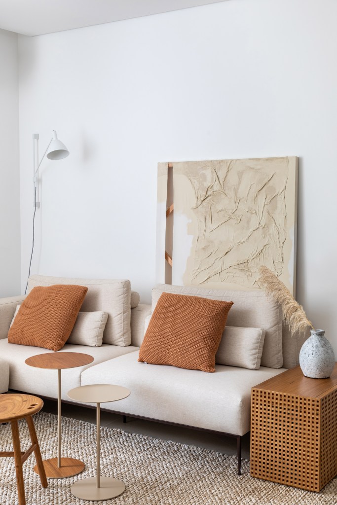 Sala de estar; sofá branco; quadro atrás do sofá; almofada marrom; mesa de centro de madeira; mesa lateral de madeira