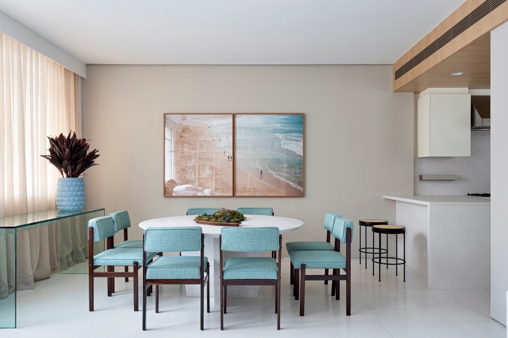 Sala de jantar em tons neutros; fotografias de praia na parede; mesa branca; cadeiras estofadas azul claro;