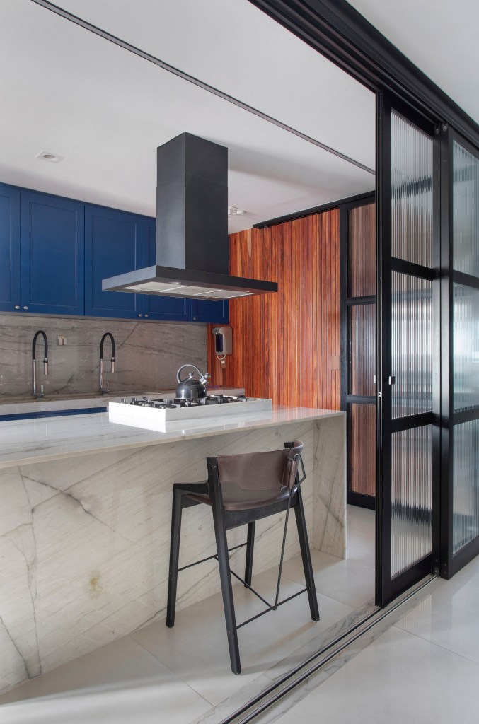 Cozinha com ilha revestida de mármore e marcenaria azul; porta de correr de vidro.