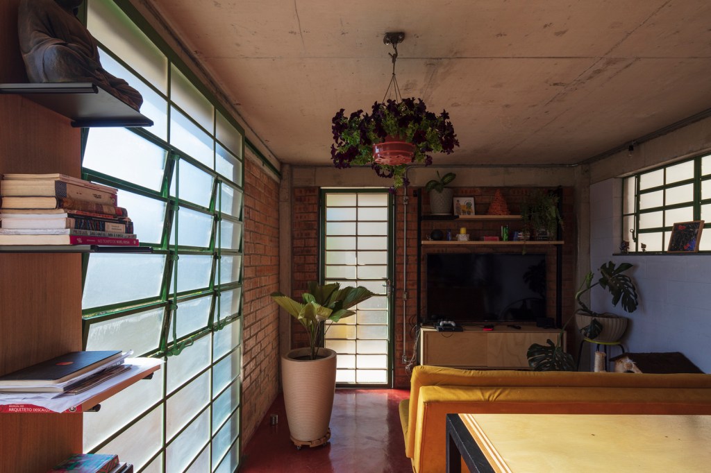 Sala de estar com janelas metálicas, sofá amarelo, planta suspensa e piso de vermelhão.