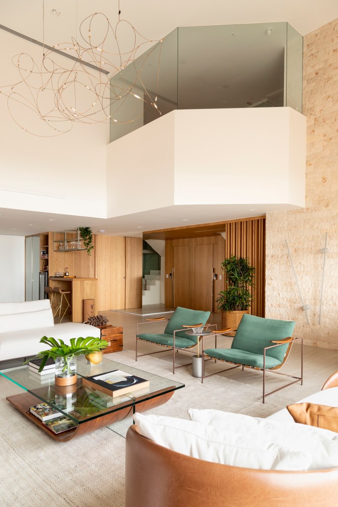 Sala de estar com pé-direito alto, sofás brancos, tapete claro, mesa de centro com tampo de vidro e poltronas verdes.