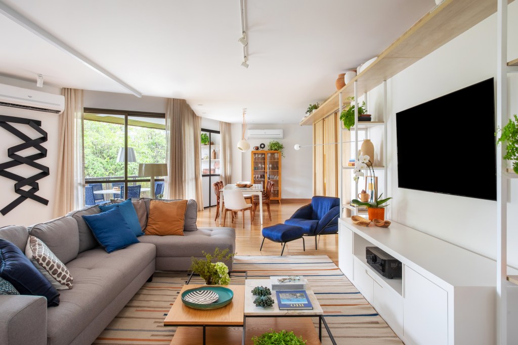 Sala de estar clara com piso em madeira, estante branca, poltrona azul, sofá cinza e tapete listrado.