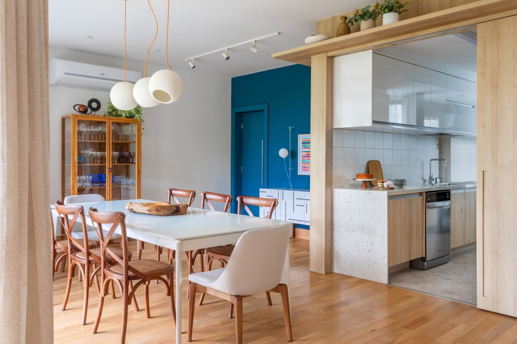 Sala de jantar integrada com cozinha; mesa branca e cadeiras de madeira; parede azul