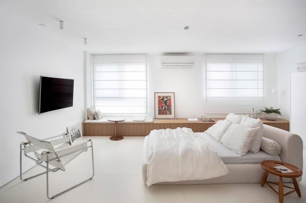 Loft branco com cama branca baixa e banco de madeira ripada