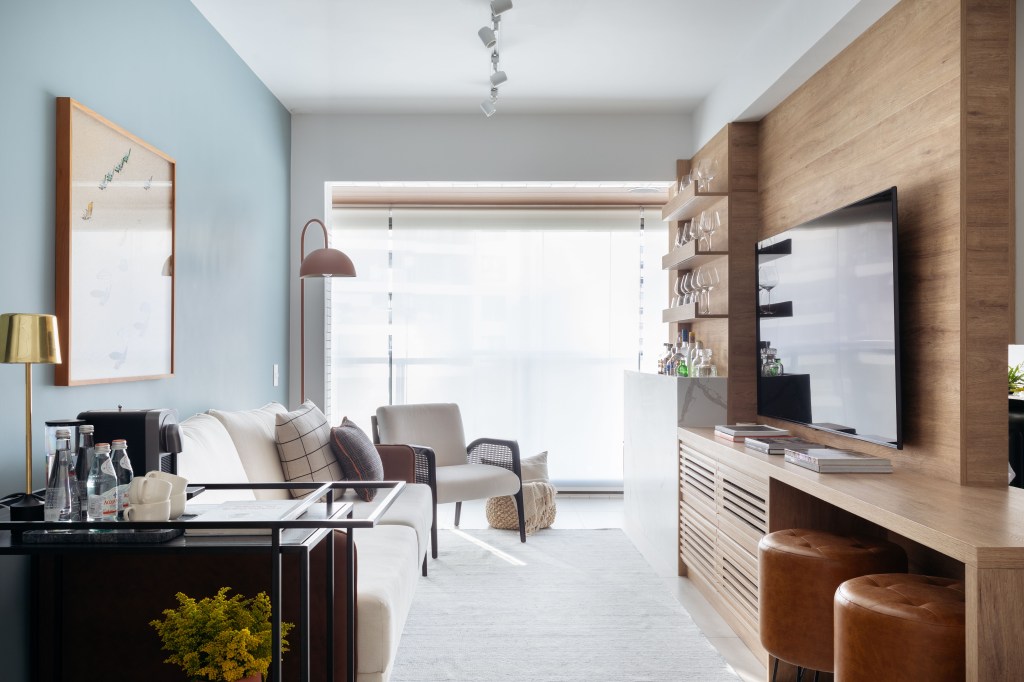 Sala de estar pequena com parede azul, sofá branco, poltrona branca e bar integrado.