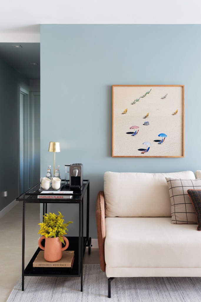 Sala de estar com parede azul, sofá branco, quadro em cima do sofá, mesa lateral metálica preta.