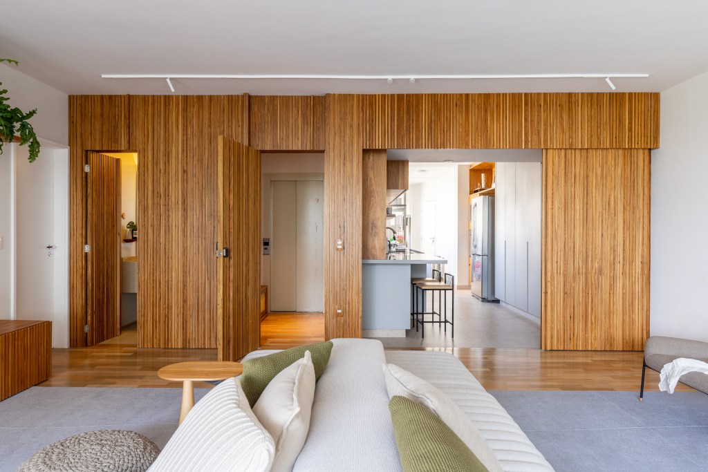 Sala de estar com sofá ilha branco; painel de madeira ripada com porta de correr para cozinha