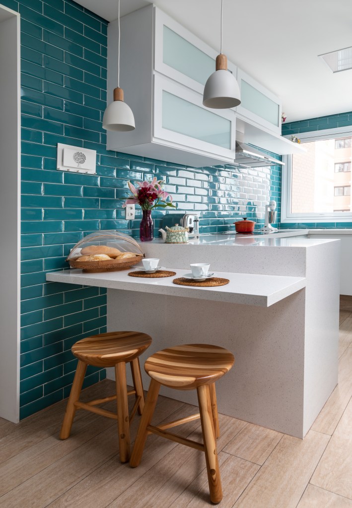 Cozinha pequena com parede de azulejos azuis e bancada pequena branca com banquetas de madeira.