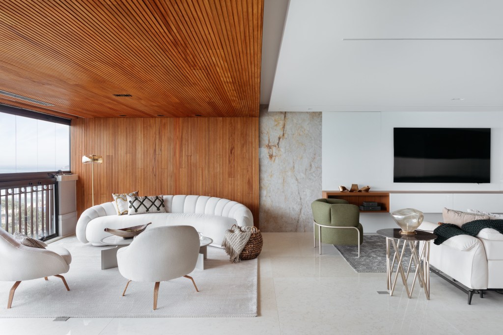 Sala de estar integrada com varanda; parede revestida de madeira e sofá curvo branco.