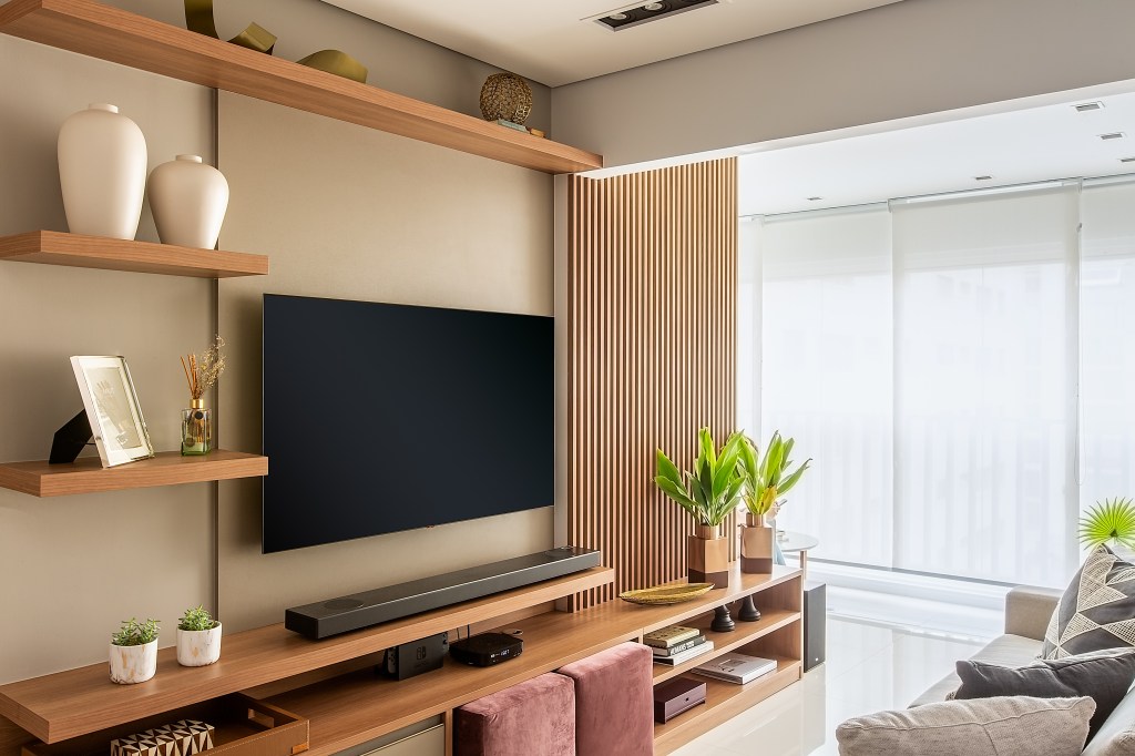 Sala de tv com painel de madeira ripada, rack de madeira e pufe na cor berinjela; varanda integrada.