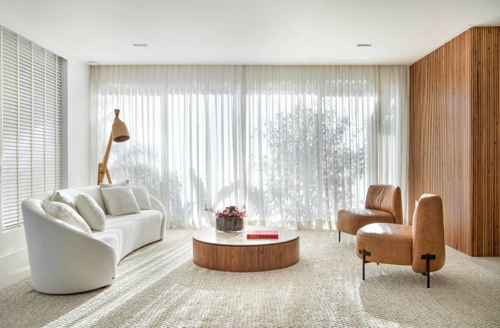Sala minimalista em tons claros; sofá branco claro curvo, tapete claro, poltrona, mesa de centro.