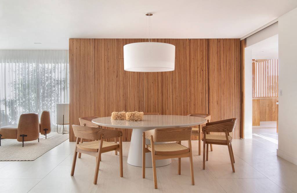 Sala de jantar minimalista com mesa redonda, luminária, cadeiras de madeira e parede revestida com madeira ripada.