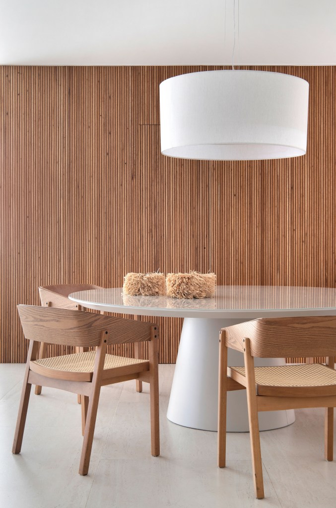 Sala de jantar minimalista com mesa redonda, luminária, cadeiras de madeira e parede revestida com madeira ripada.