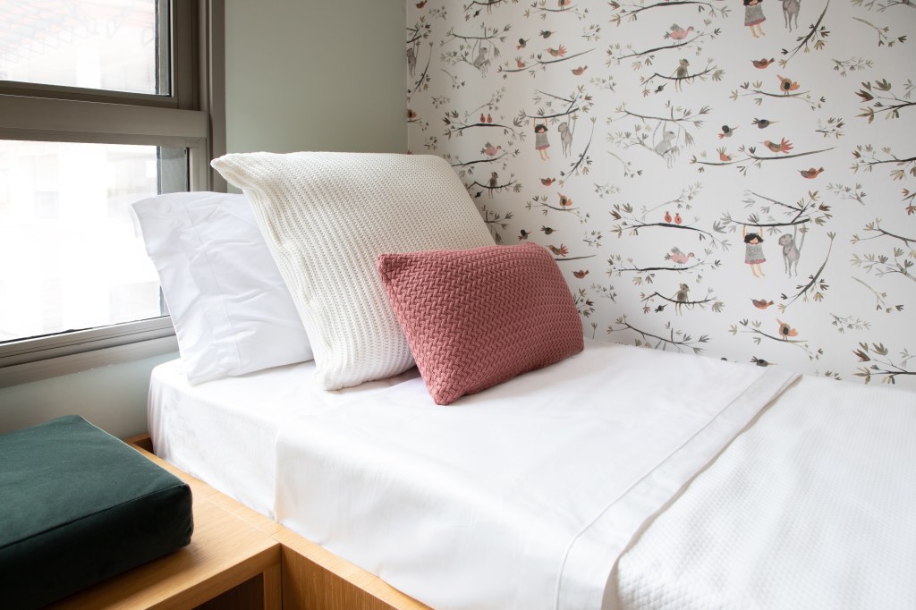 Quarto com cama de solteiro, lençol branco, três travesseiros e papel de parede.