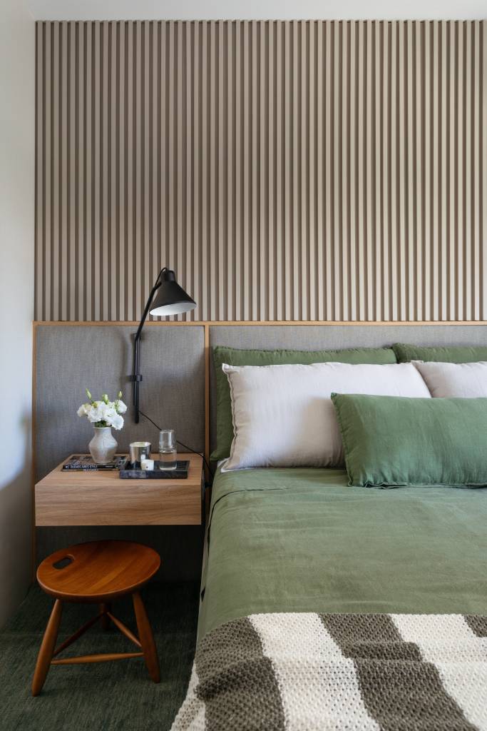 Quarto com cama de casal e roupa de cama verde; cabeceira estofada e parede revestida com painel ripado.