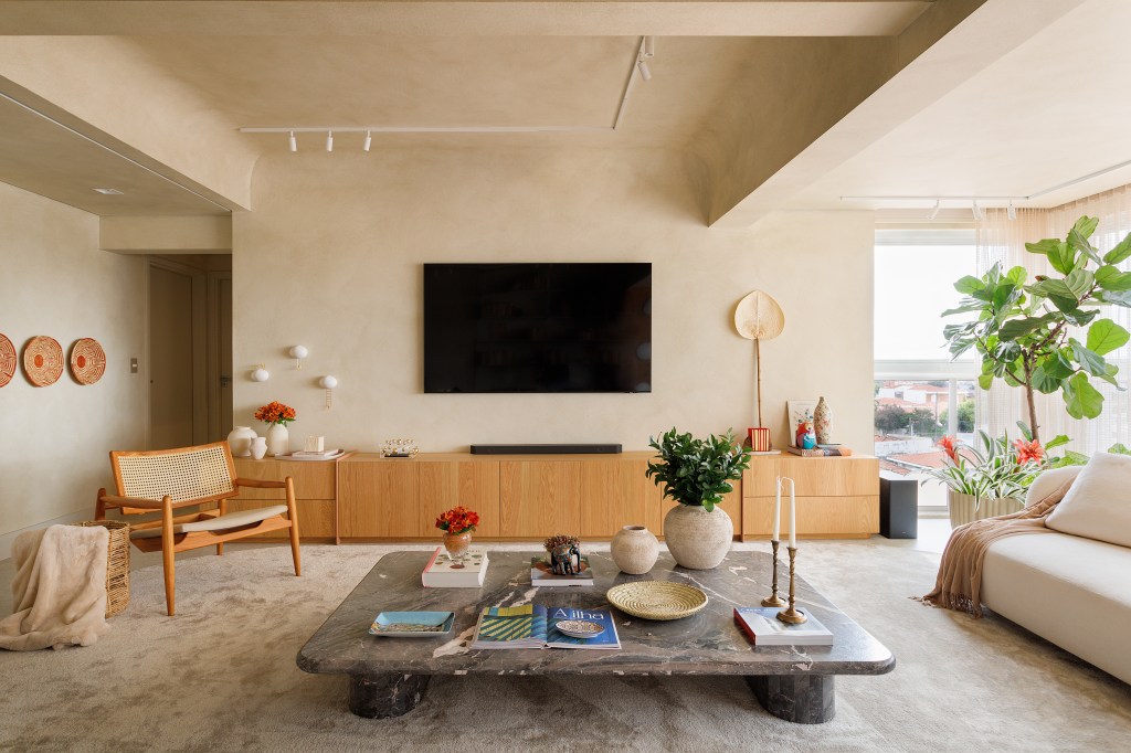 Sala de tv em tons terrosos claros com mesa de centro, sofá branco e rack de madeira.