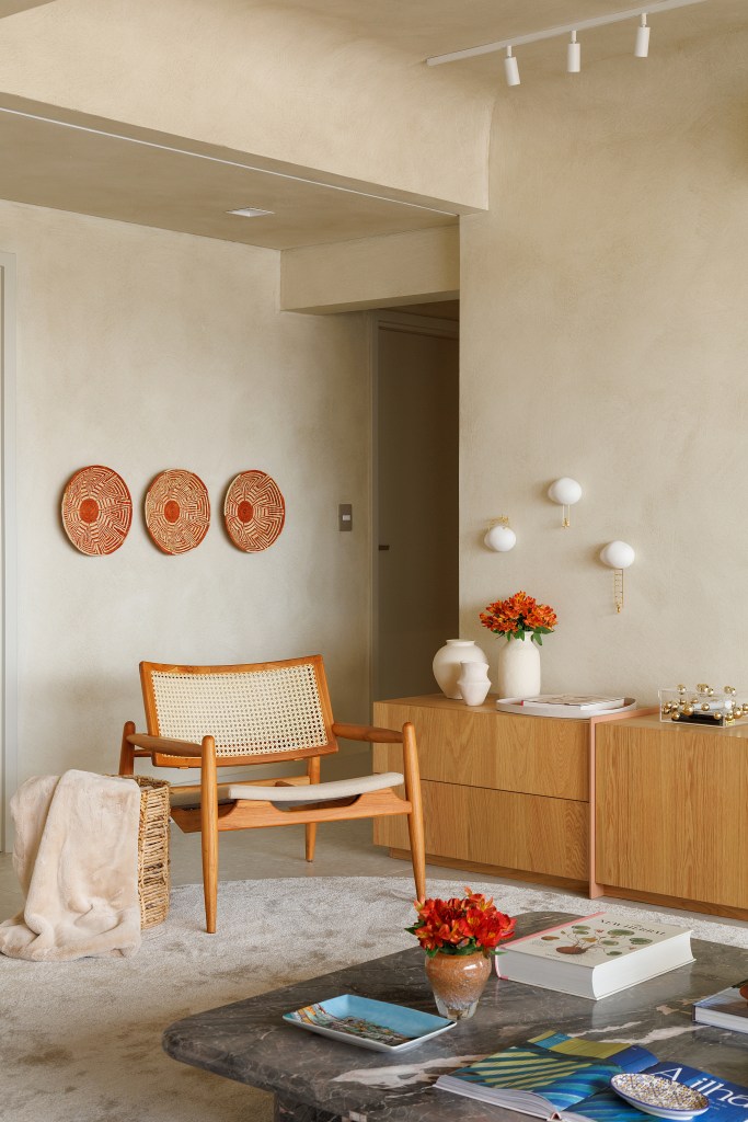 Sala de estar com paredes revestidas na cor areia; poltrona e rack de madeira.