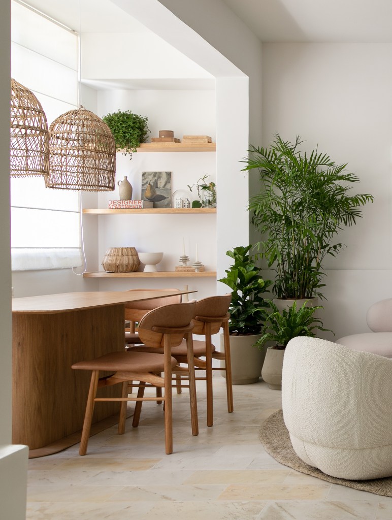 Sala de jantar em tons claros com mesa e cadeira de madeira; prateleiras de madeira com vasos e plantas.