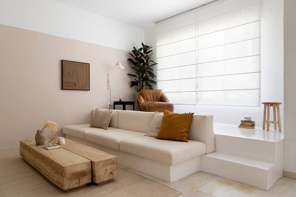 Sala de tv em tons claros com rack branco, sofá branco e mesa de centro de madeira rústica; degrau embaixo de janela para cantinho de leitura.