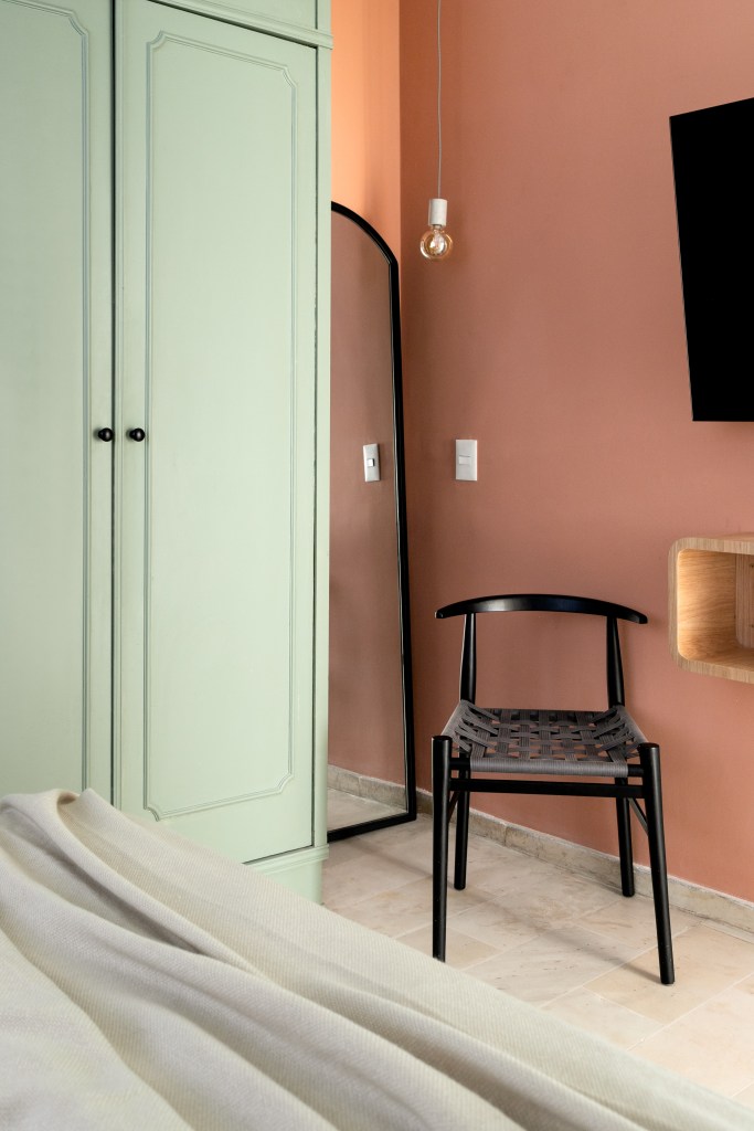 Quarto com armário verde, espelho de piso, cadeira preta e parede rosa terrosa.