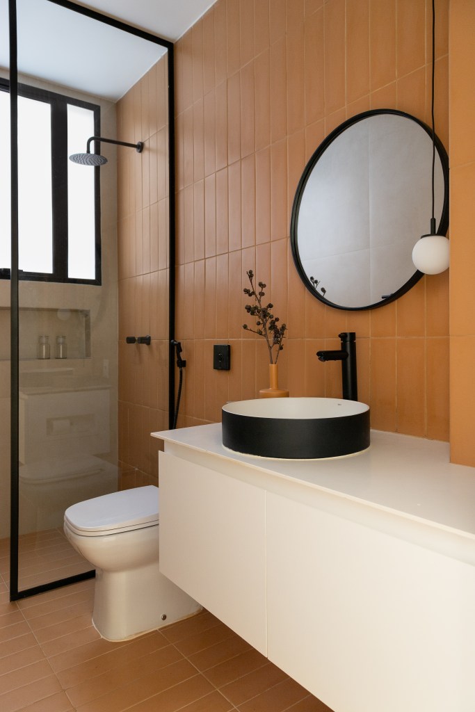 Banheiro com parede revestida de azulejos laranja, box de vidro e espelho redondo.