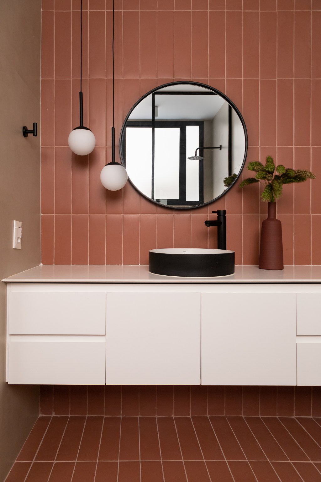Banheiro com parede de azulejos rosa terroso, armário branco e espelho redondo.