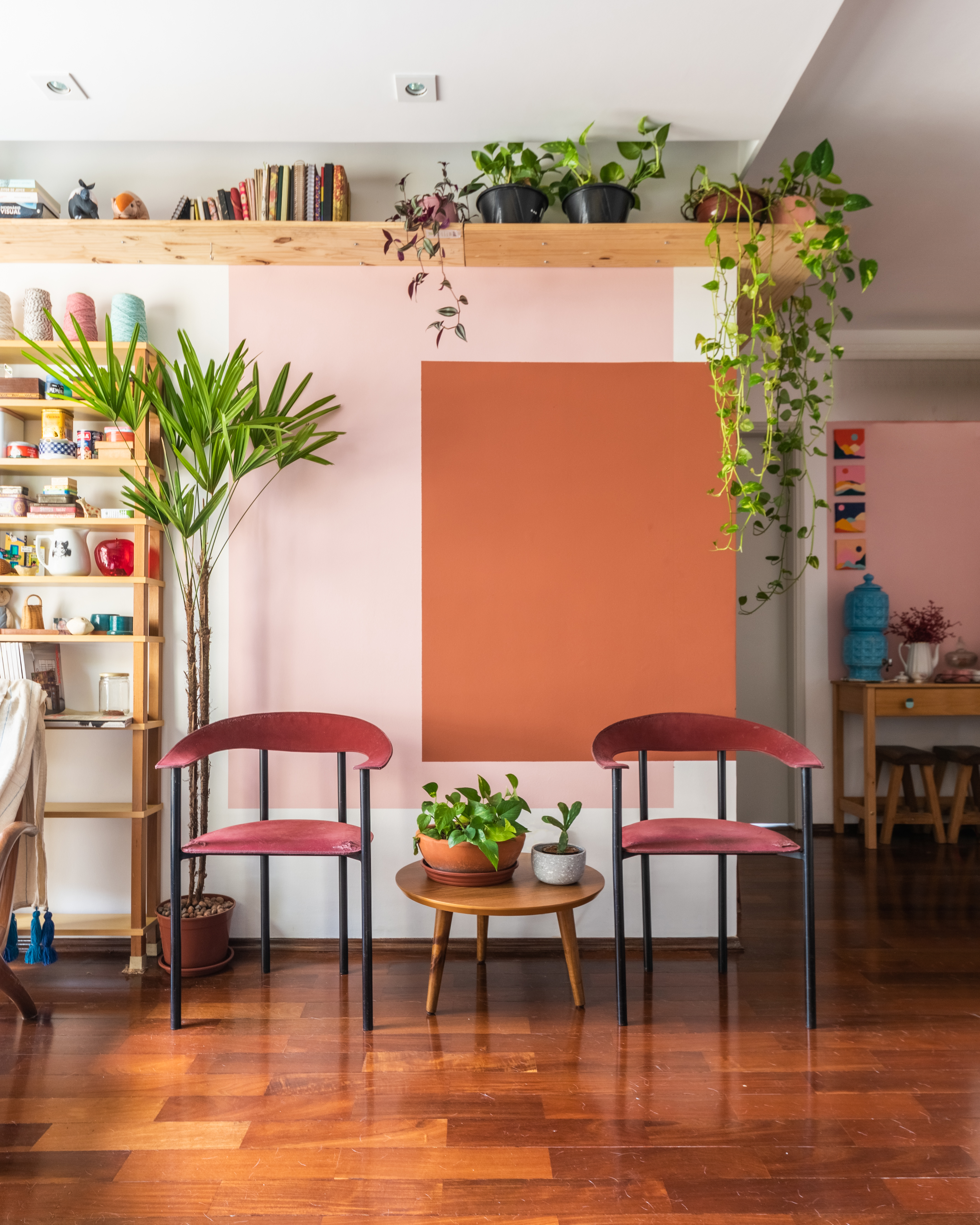 Sala de estar com parede rosa e pintura geométrica terracota, plantas suspensas e palmeira.