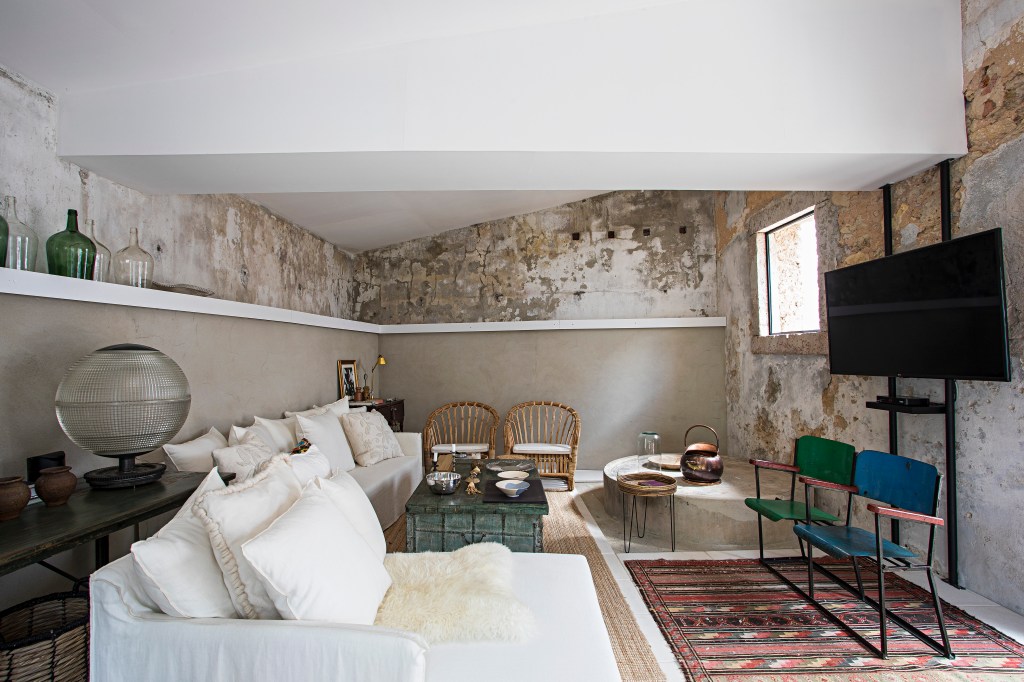 Sala de estar rústica com sofá branco, paredes expostas, mesa de centro e tapete colorido.