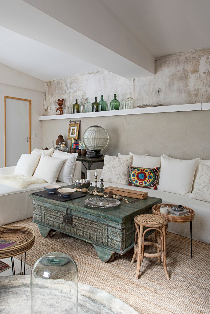 Sala de estar rústica com sofás brancos, mesa de centro baú, parede exposta e prateleira com garrafas