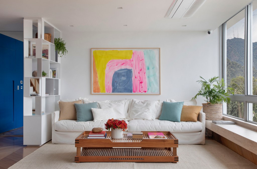 Sala de estar com sofá branco, tapete bege, poltrona e quadro colorido.
