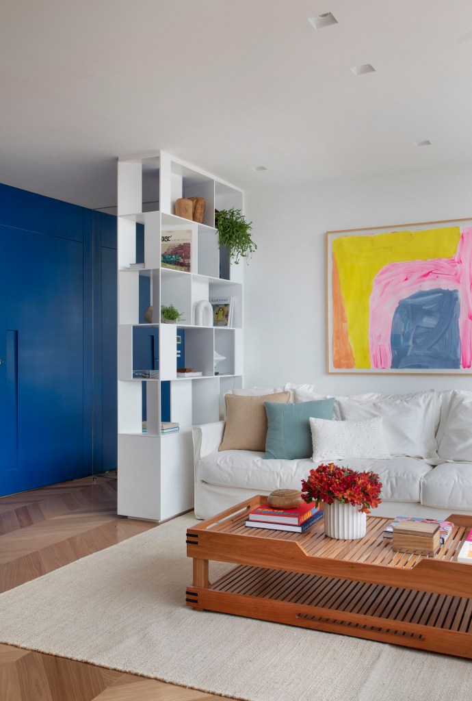 Sala de estar com sofá branco, tapete bege, poltrona e quadro colorido, parede azul e estante branca vazada.