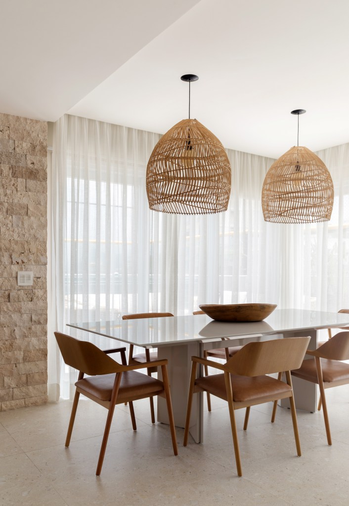 Sala de jantar com mesa de jantar, cadeiras de madeira, luminária de palhinha e cortina branca.