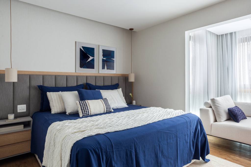 Quarto com cama de casal e roupa de cama azul, cabeceira acolchoada cinza.