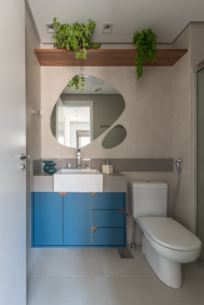 Banheiro com espelho em forma orgânica, armário azul e plantas.