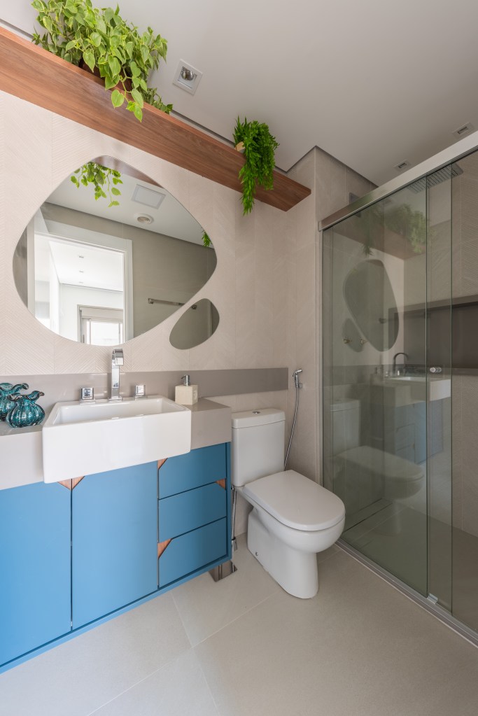 Banheiro com espelho em forma orgânica, armário azul e plantas.