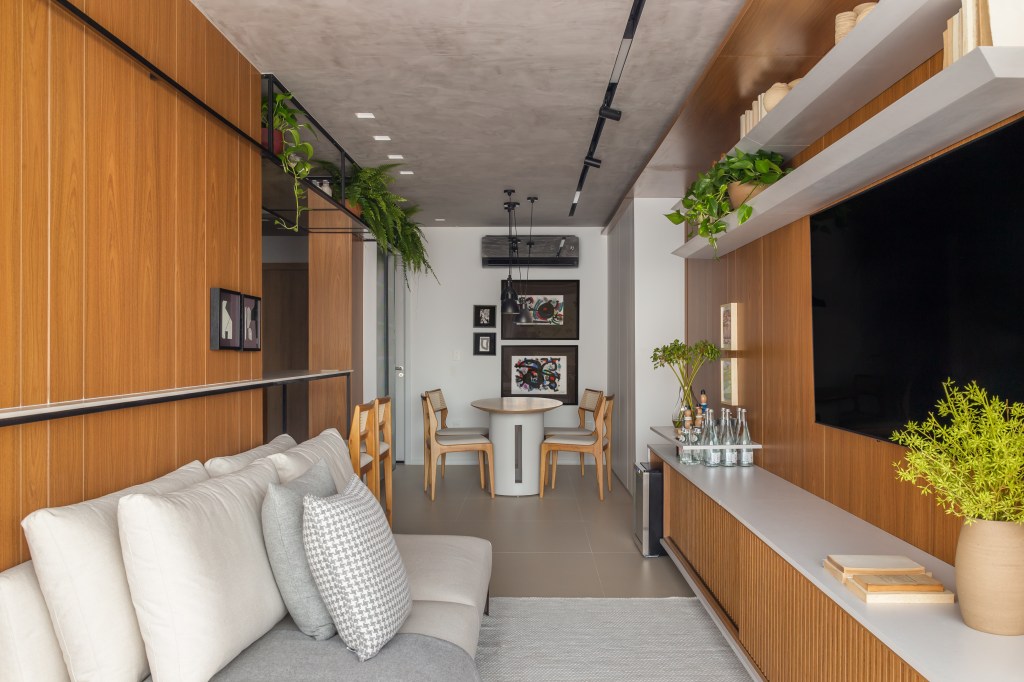 Sala de estar integrada com varanda, cozinha e estar. Sala estreita com sofá cinza e parede revestida de madeira.